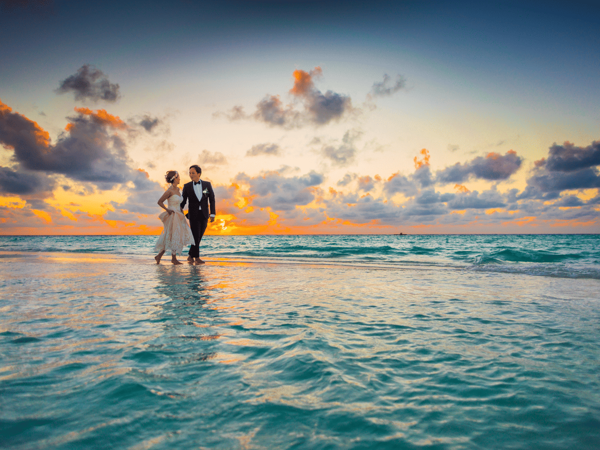 A wedding picture of a couple walking on the shallow seashore in the evening with a sunset view in the backdrop.