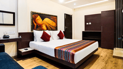 A modern hotel room with wooden flooring, a double bed featuring white linens and red cushions, a vibrant wall painting above the headboard, a dark sofa, and a functional work desk - Mount Hotels