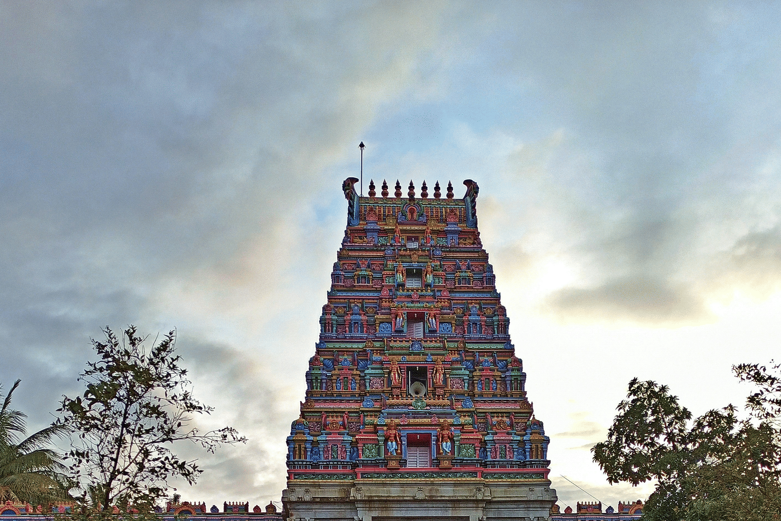 An ancient temple gopuram with a paved path leading towards the structure under a partially cloudy sky.