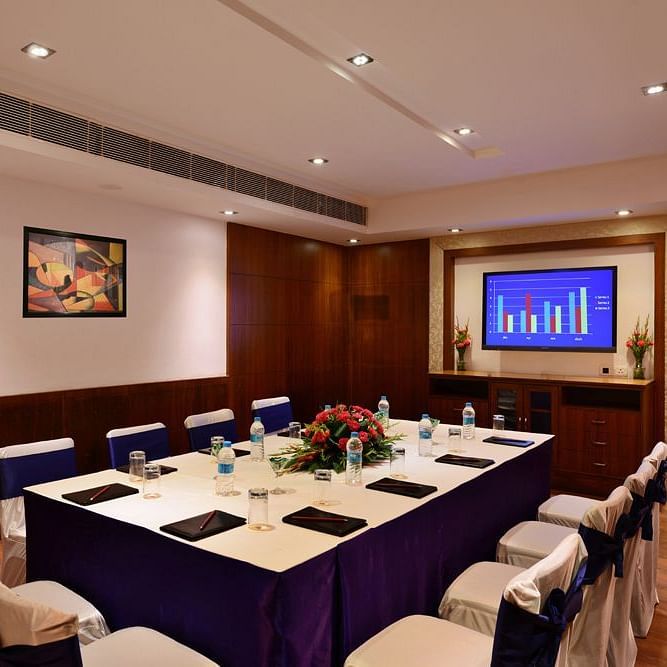 Board room with royal blue seating arrangement, bright lighting, flat screen TV, and wooden flooring.
