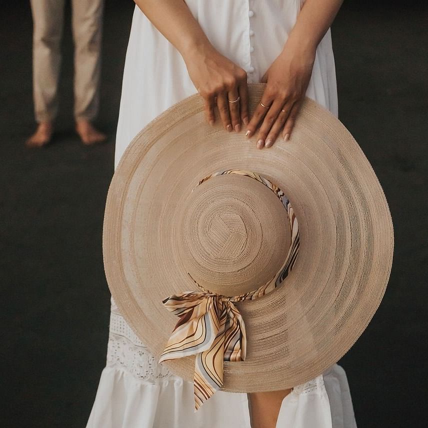 The image shows a person holding a large straw hat with a ribbon, wearing a white dress, while a person in the background is partially visible.