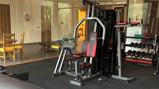 A gym interior with a treadmill in the foreground and various strength training equipment, including a multi-gym station, visible in the background; the space features glass walls and a seating area outside the workout zone - Silver Sand Beach Resort, Have