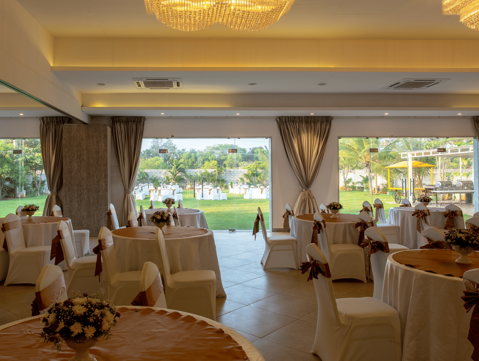 An indoor banquet hall with round tables and chairs draped in white with brown sashes, set for an event - Grande Bay Resort & Spa, Mamallapuram