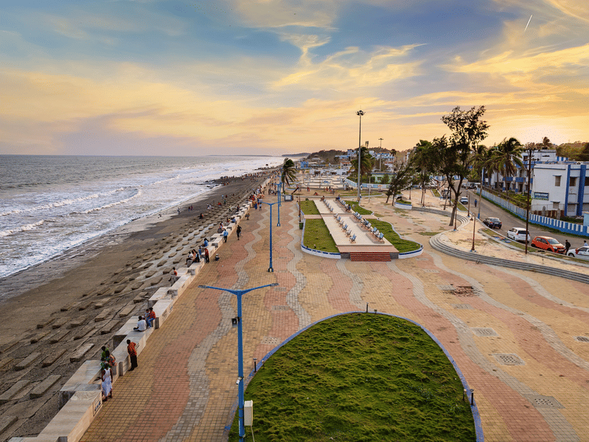 People stroll along the paved sea-facing promenade and sandy shores of Digha Beach during a warm, vibrant sunset over the ocean waters.