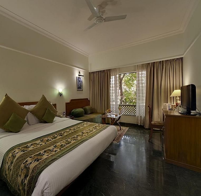 A comfortable hotel room at Renest Shraddha Inn features a bed with decorative pillows and a patterned runner, a seating area, a television on a stand, and large windows with curtains - Renest Shraddha Inn, Shirdi
