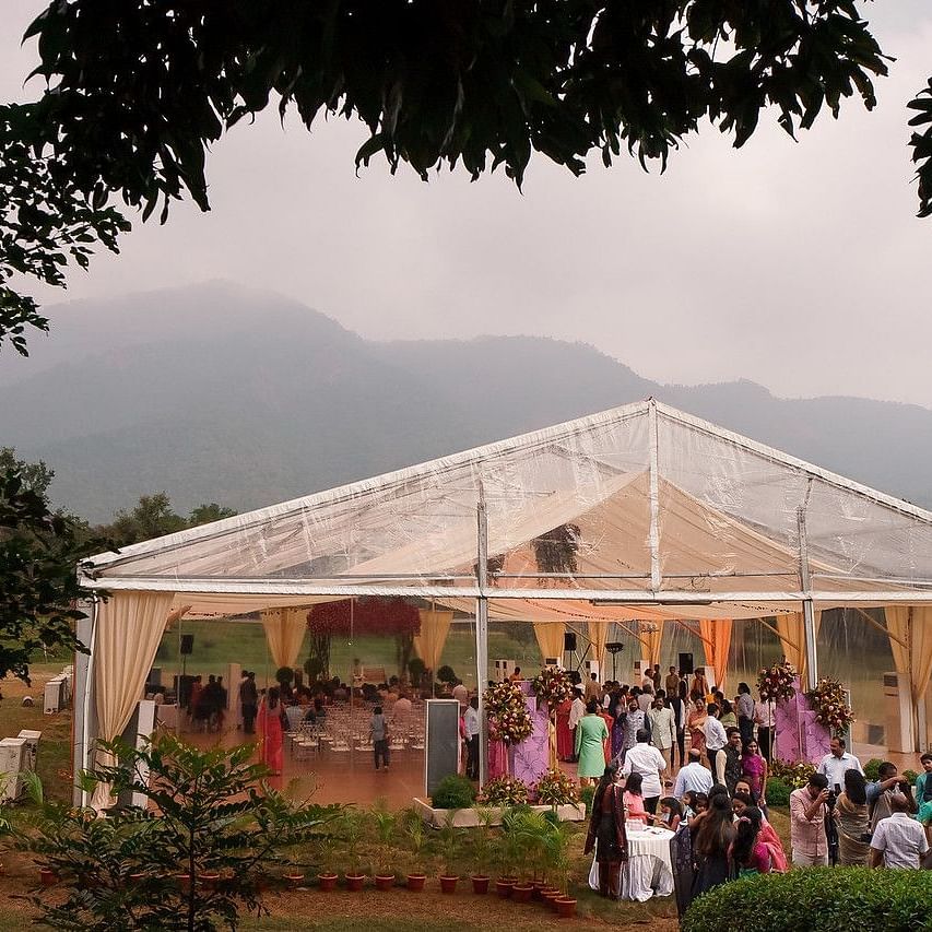 An outdoor event with guests gathered near a clear marquee tent, surrounded by nature and mountains in the background, creating a scenic and festive atmosphere at Black Thunder, Coimbatore