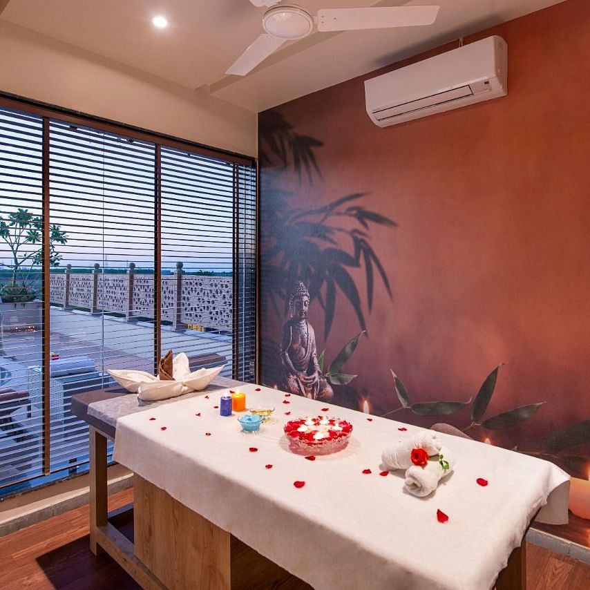 A serene spa treatment room featuring a massage table draped with a white sheet and adorned with red rose petals and decorative items, set against a warm-toned wall with a subtle mural and with natural light coming through large windows overlooking an outd