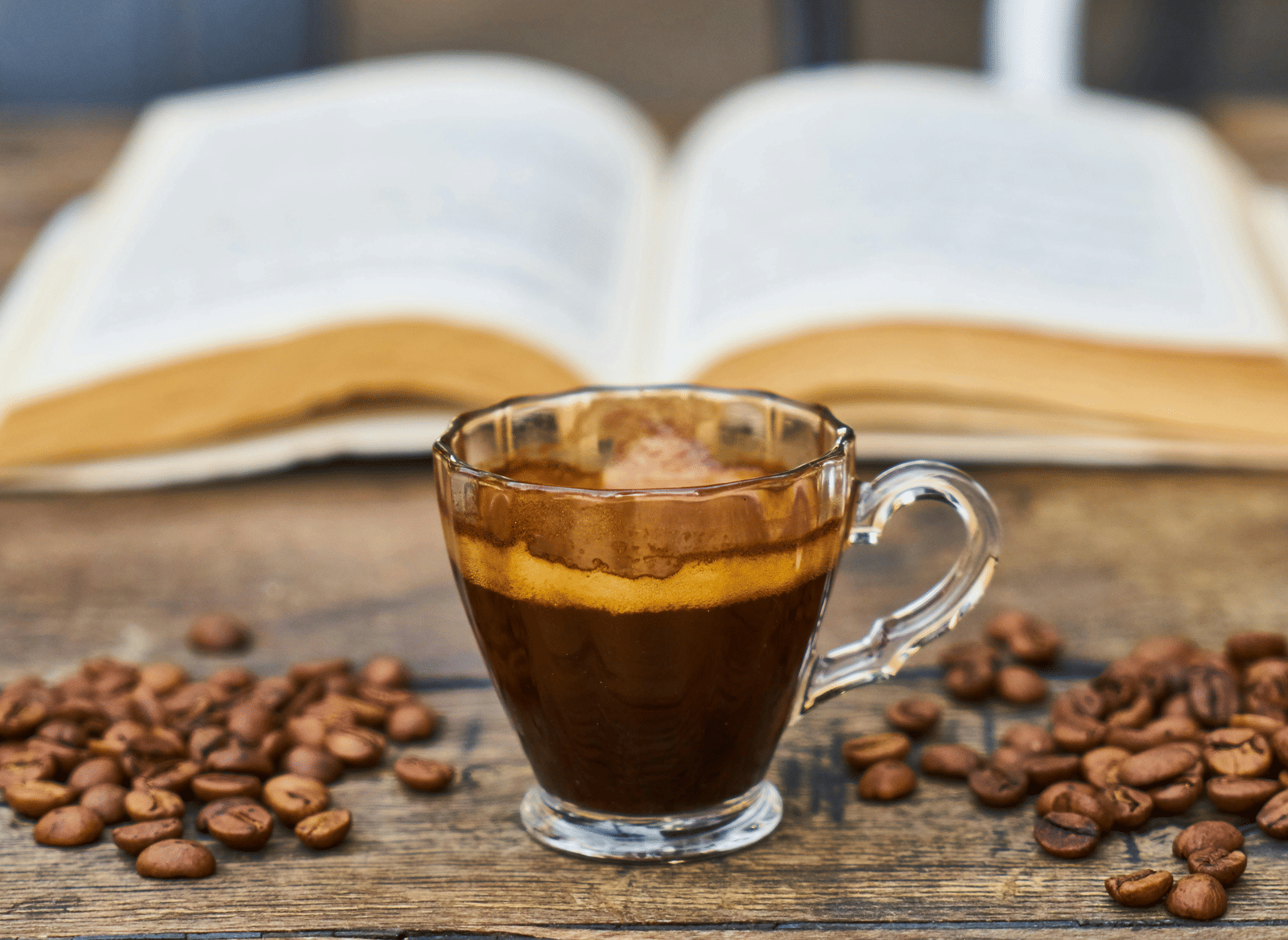 A cup of coffee placed on a wooden table beside an open book and coffee beans.