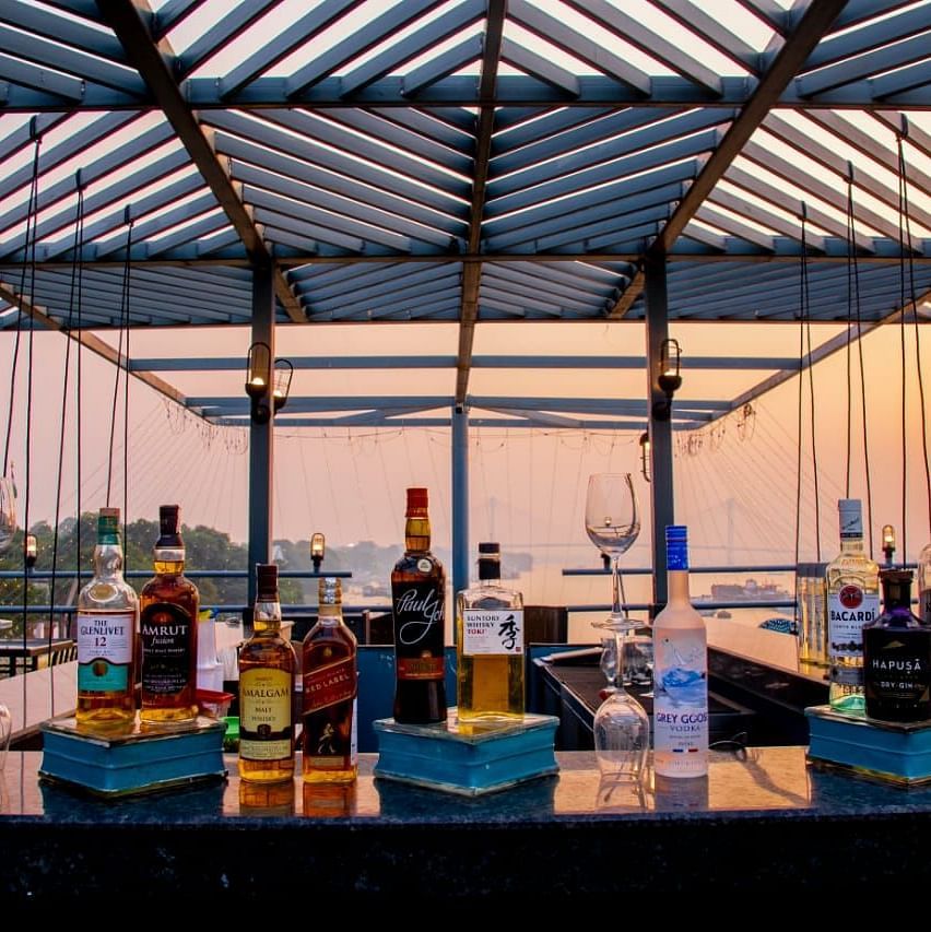 An outdoor bar on a rooftop with bottles of liquor lined up against a backdrop of sunset skies and a river view.