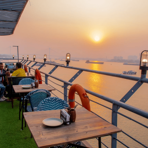 People sitting at tables with a sunset view over a river, seen from a rooftop bar with a grassy floor and metal railing.