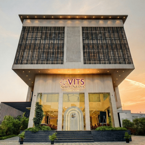 Facade of VITS Select Nashik Hotels & Resorts, one of the best hotels in Nashik for family stays, under a vibrant sky with the well-lit exteriors surrounded by bushes and an open area surrounding the hotel.