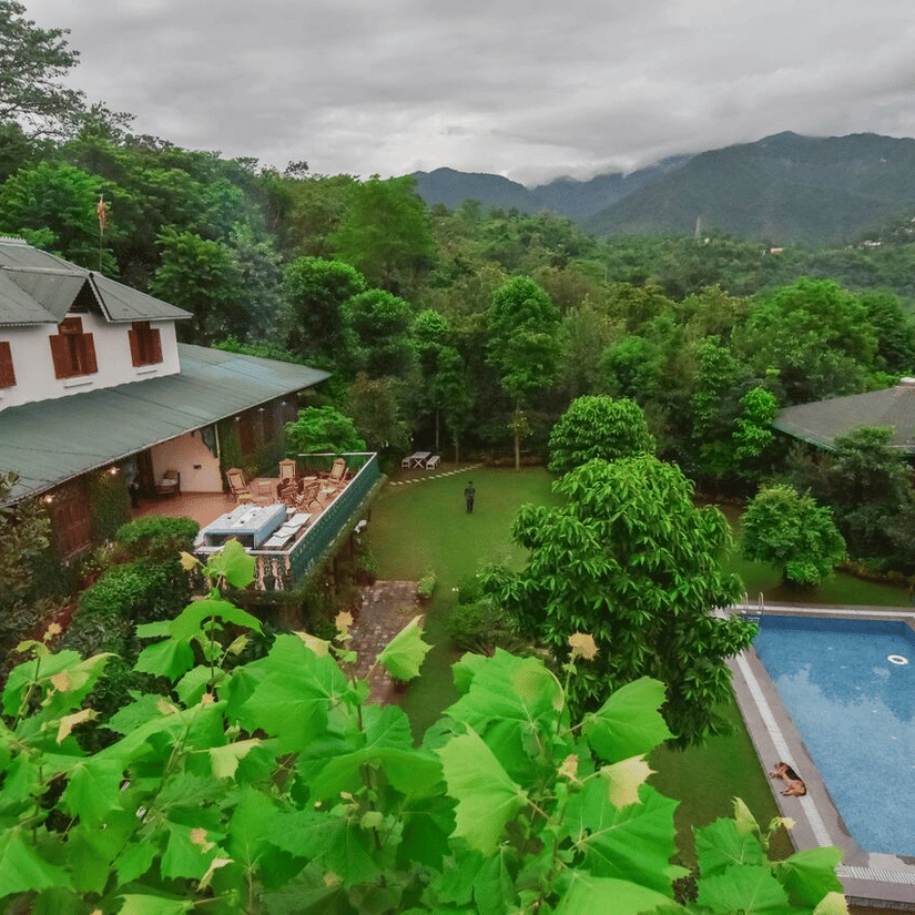 Facade of Shaheen Bagh - A Luxury Boutique Resort & Spa in Dehradun, surrounded by trees with a swimming pool next to the lawn area and mountain ranges in the background.