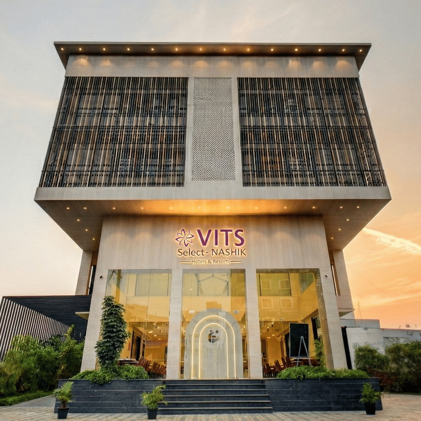 Facade of VITS Select Nashik Hotels & Resorts, one of the best hotels in Nashik for family stays, under a vibrant sky with the well-lit exteriors surrounded by bushes and an open area surrounding the hotel.