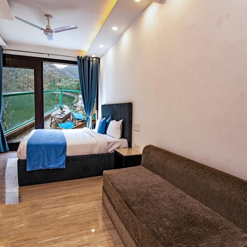 A bedroom with a large bed, sofa, wooden flooring, and full-length windows overlooking greenery.