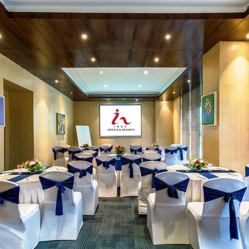 Meeting room at Inde Reva Resorts Rishhikesh, with white chairs tied with navy blue sashes, round tables decorated with flower arrangements, and a projector screen at the front.