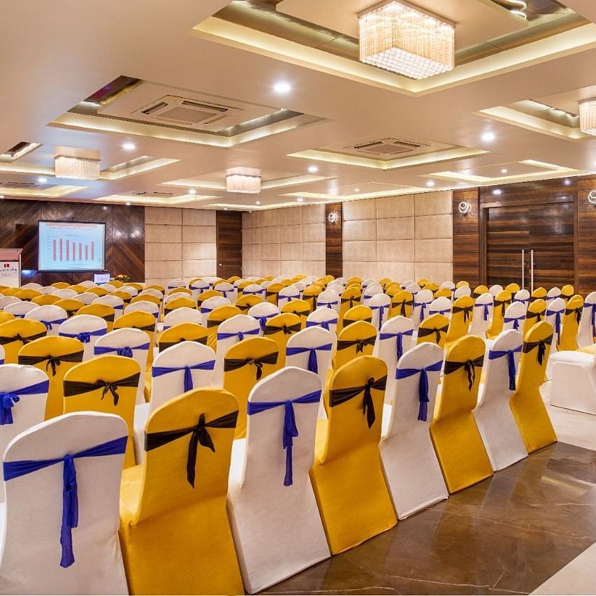 A large banquet hall at Lords Inn Porbandar, is arranged with rows of chairs covered in white and yellow fabric for an event.