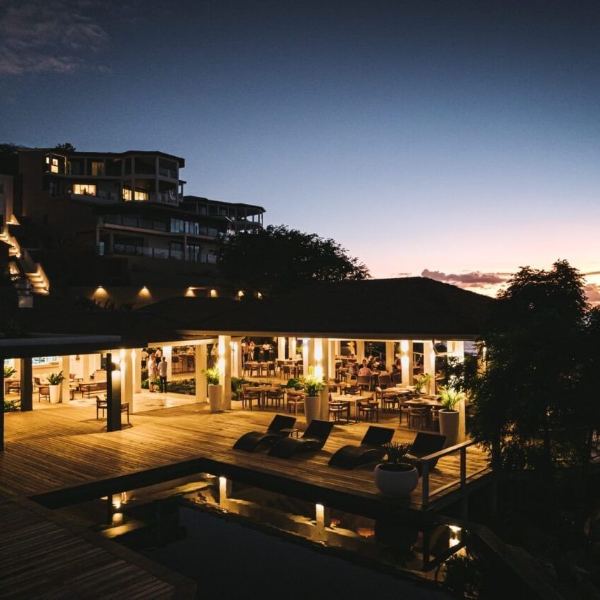 A coastal resort at dusk, with illuminated buildings, an outdoor patio, and a pool, under a twilight sky.