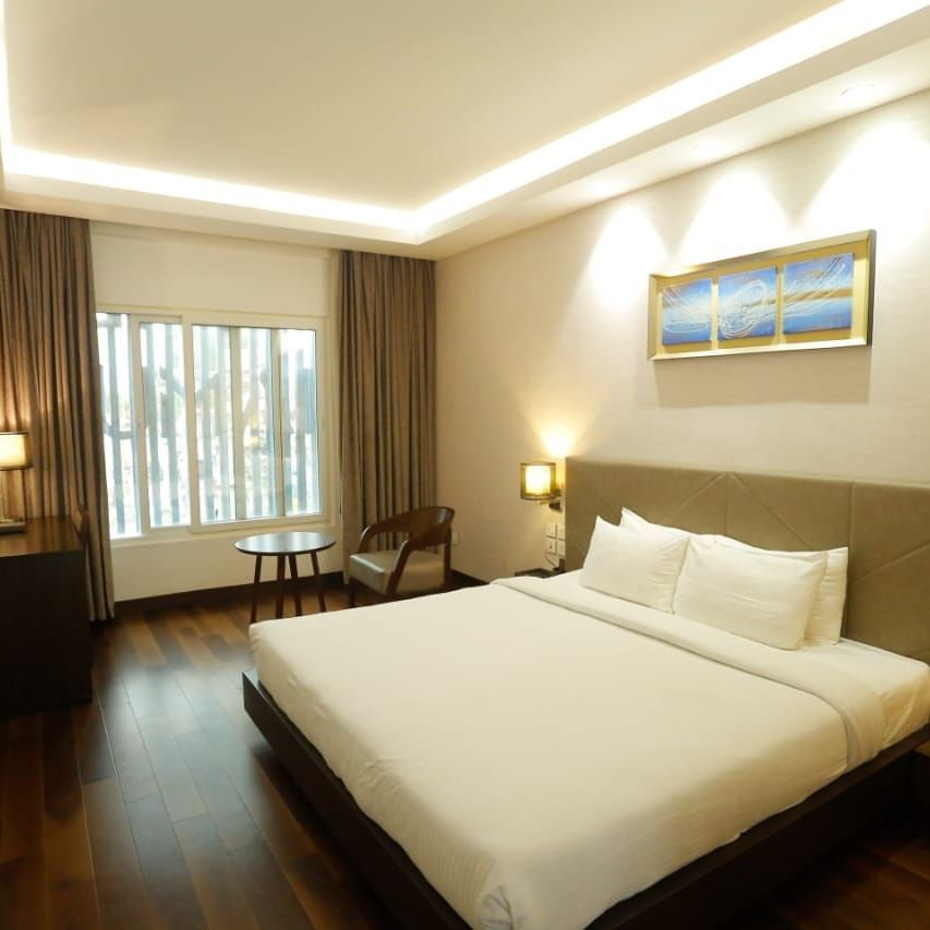 Spacious room with a large double bed, bedside lamps, wall art above the headboard, and a window with curtains pulled to the side at The Hamuse Hotel, Kodaikanal.