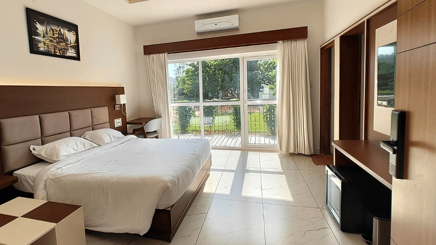 A hotel bedroom featuring a double bed, wooden furniture, a wall-mounted picture frame, and large sliding doors looking out towards greenery | Lords Sahyadri Resort, Shivamogga