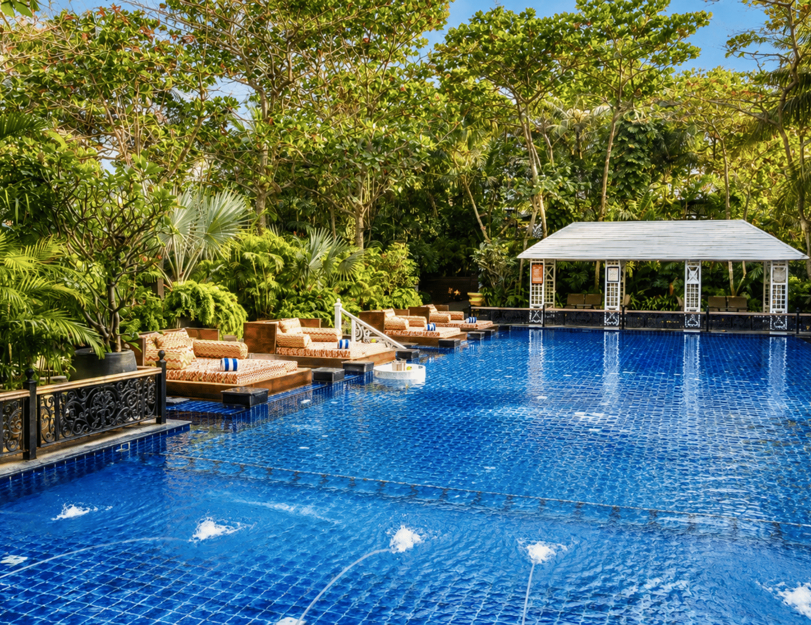 Outdoor swimming pool with blue tiling, cushioned daybeds, a white poolside pavilion and surrounding greenery at MAYFAIR Palm Beach Resort, Gopalpur-on-Sea.