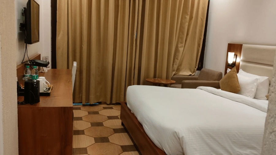 A standard room at Pride Ashiyana Resort Mussoorie with a bed, TV, desk, chair, and beige curtains.