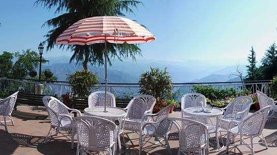  A serene outdoor dining area at Hotel Mount View featuring white wicker tables and chairs under colourful umbrellas, overlooking a vast body of water and lush green trees against a clear blue sky. 