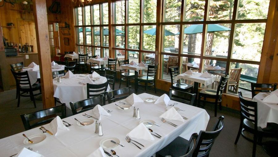 A dining area with multiple tables set for meals, featuring large windows looking out to trees