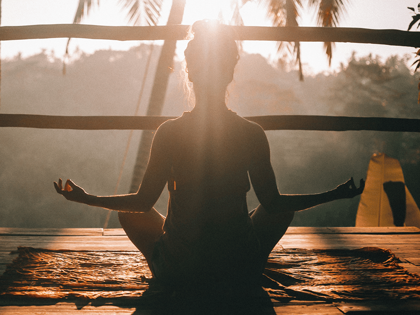 A woman meditating cross-legged on a wooden deck at sunrise, silhouetted against palm trees