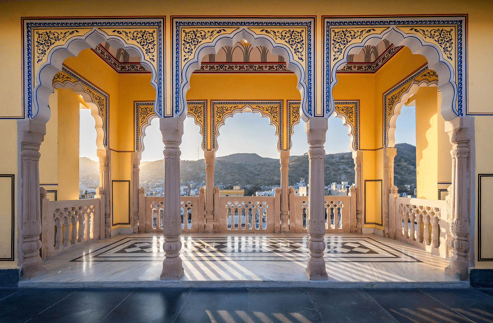a balcony at the rooftop with yellow walls and intricate paintings on the pillars with blue colour with the sun rays brightening up the area - Brij Castle, Jaipur