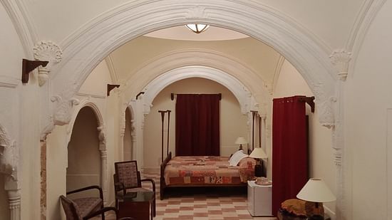 Arched doorways lead through the white interiors of Tijara Fort-Palace - 19th Century, Alwar, revealing a bedroom with a patterned bed and seating.