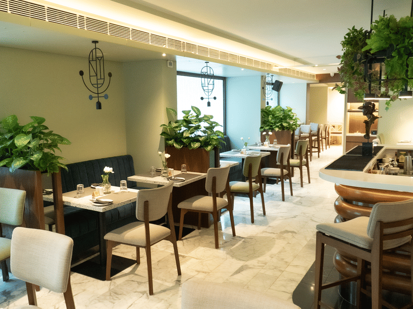 A serene dining area at the Koi Garden of Hotel Bawa International in Vile Parle featuring neatly arranged tables, upholstered chairs, indoor plants, and soft ambient lighting creating a calm and elegant setting.