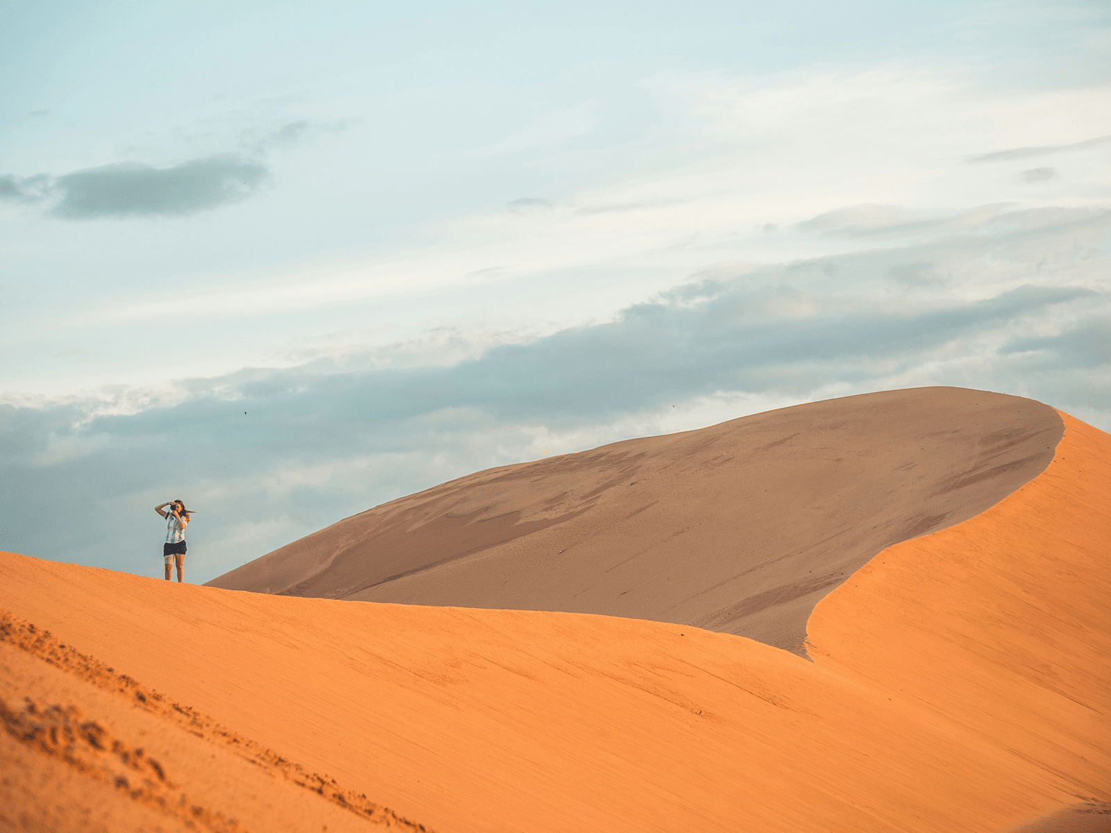 An overview of a desert in mui ne with high red dunes and a woman standing on top in view, one of the best Phan Thiet Tourist Attractions.