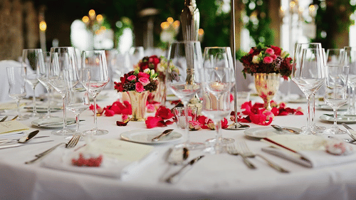 Close-up view of a formal dinner table set for an event, featuring multiple wine glasses, silverware, plates, and floral centrepieces with scattered rose petals.