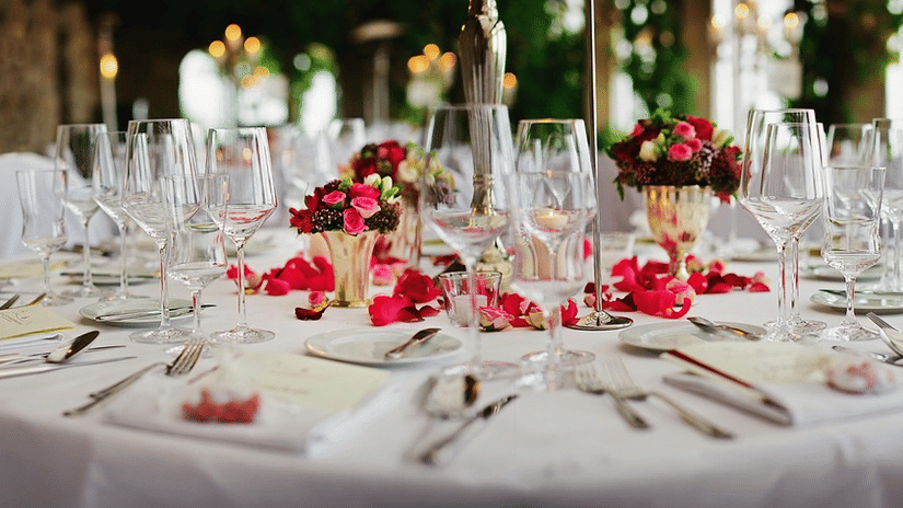 Close-up view of a formal dinner table set for an event, featuring multiple wine glasses, silverware, plates, and floral centrepieces with scattered rose petals.