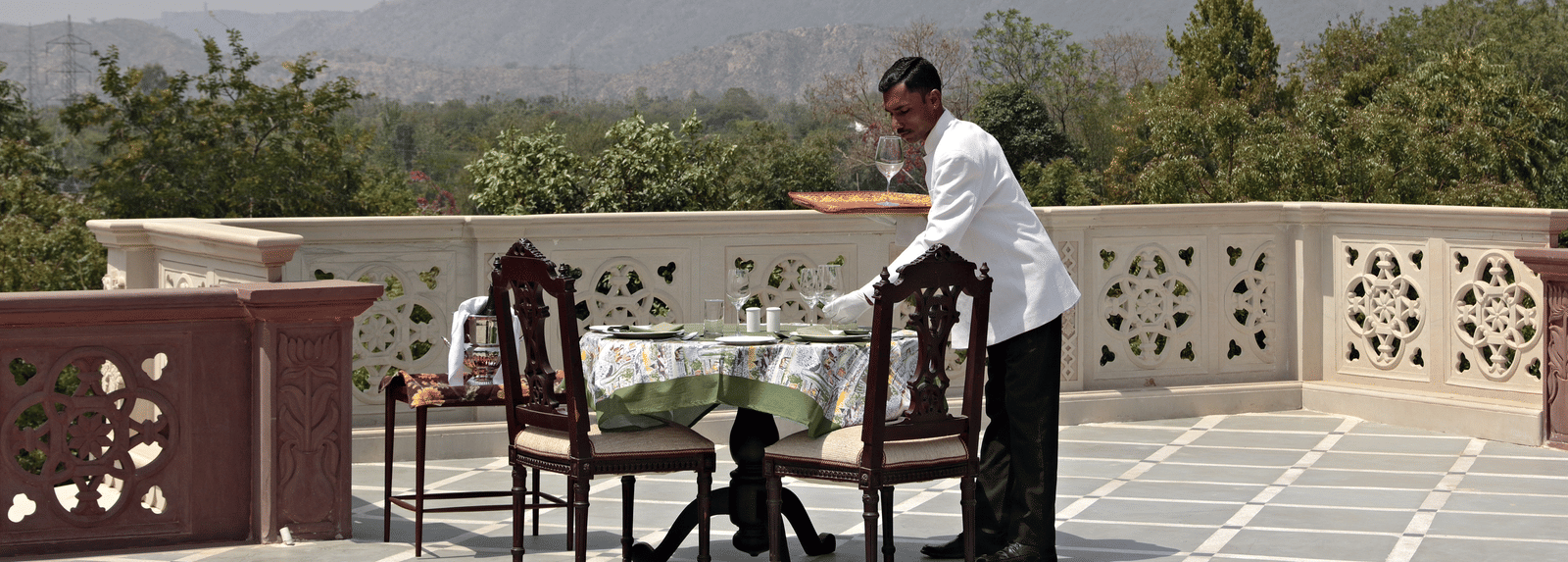 A waiter setting up a table and chairs on a balcony with a scenic mountain view - Khas Bagh, Jaipur.