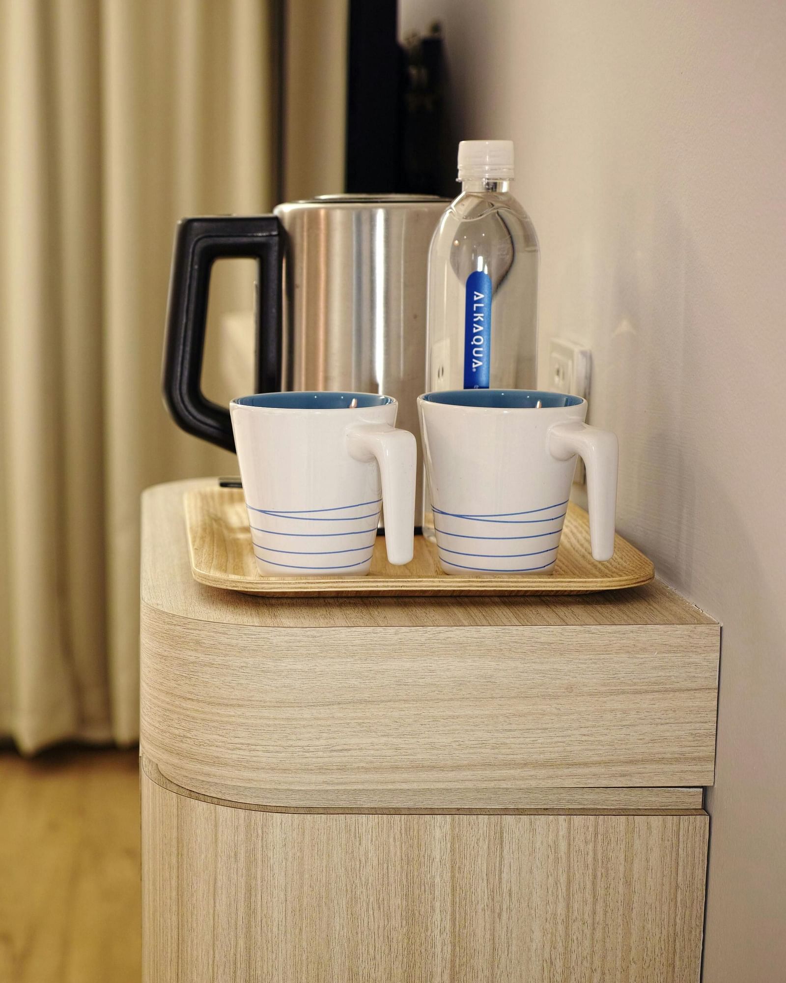 A hotel room bedside table holds mugs, a kettle, and bottled water next to a neatly made bed.