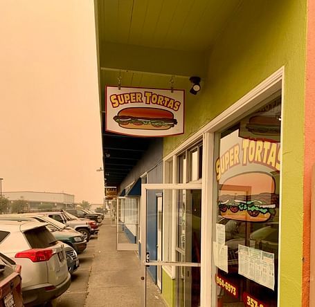 A Super Tortas restaurant with a bright green exterior features a hanging sign and window decals with a cartoon-style sandwich graphic. A row of parked cars lines the side of the building.