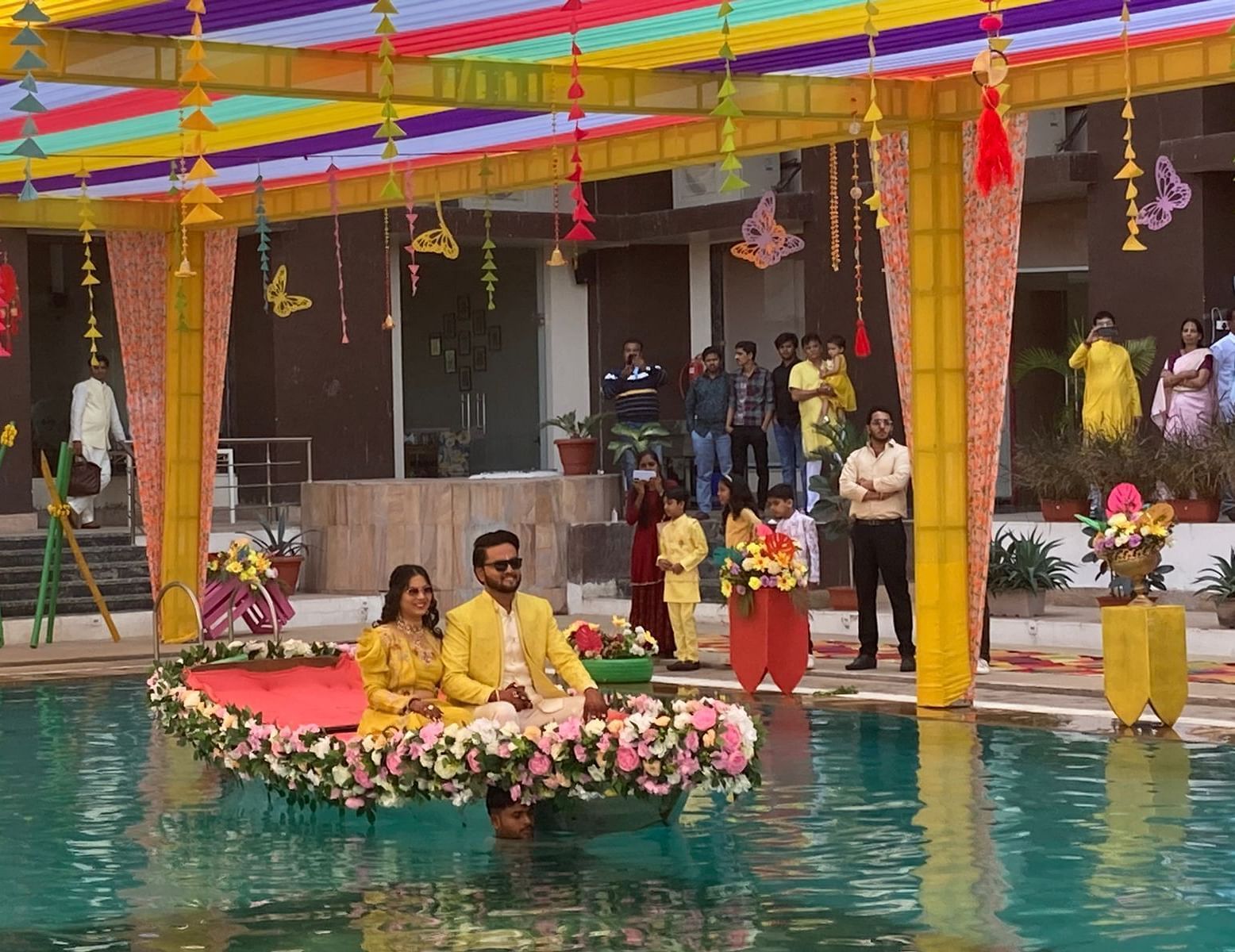 A decorated platform floating in a swimming pool, set up for a celebration. The platform is adorned with flowers and features two people sitting on it, dressed in yellow traditional clothing - Bamboo Saa Mulberry Resort, Pushkar
