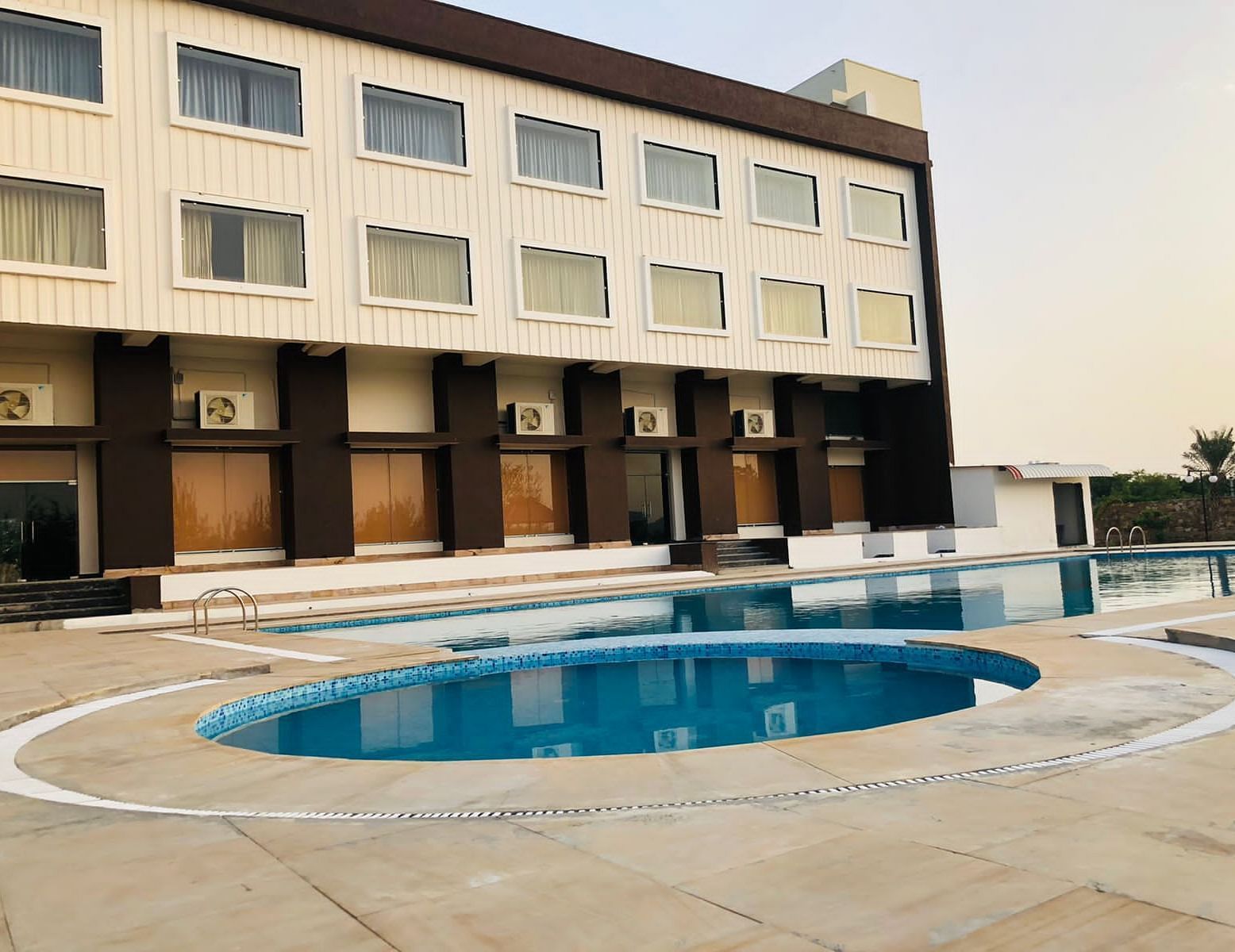The exterior of the Bamboo Saa Mulberry Resort, Pushkar, featuring a multi-story building with a light-coloured facade with a swimming pool is visible in the foreground.