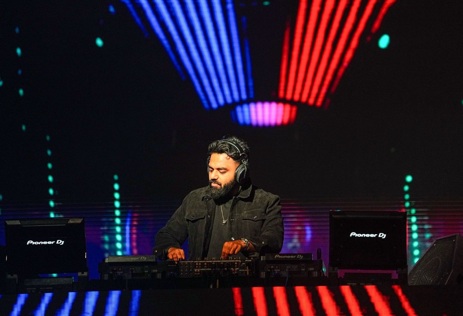 A DJ or electronic music artist performs on a stage with prominent vertical lights. The performer is silhouetted against the bright illumination and appears to be operating electronic equipment.