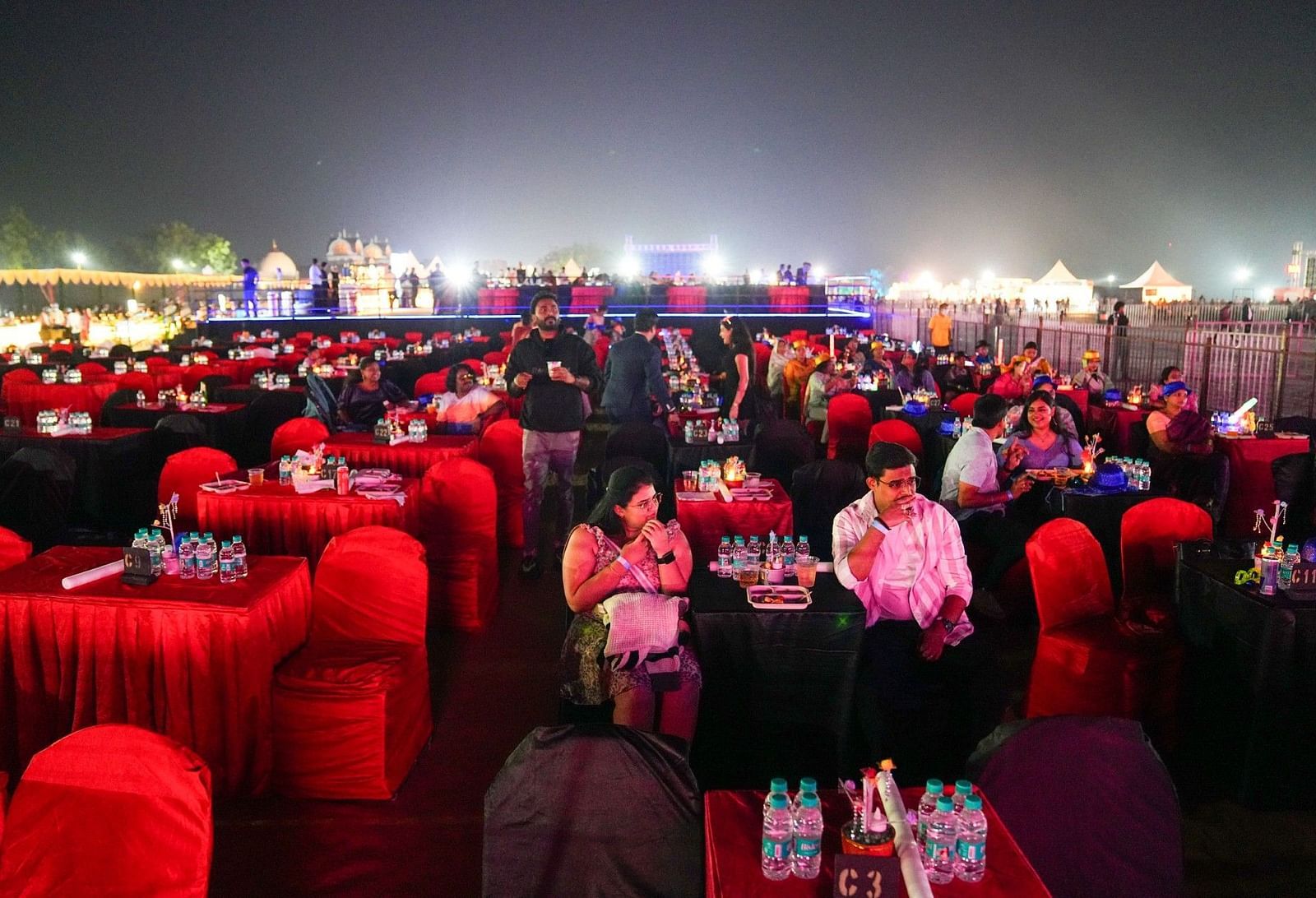 A large crowd of people, many with head coverings, are gathered outdoors at night. Bright lights illuminate the scene, suggesting a festival, concert, or large public gathering.