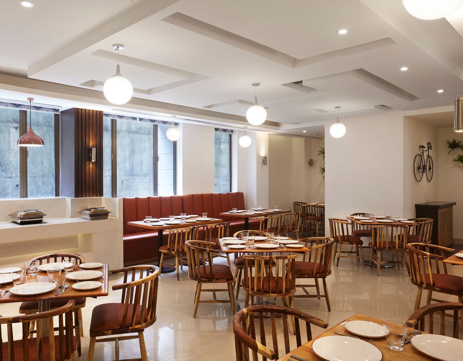 A spacious, modern dining hall at Monday Hotels Mahape, Navi Mumbai, with multiple wooden tables, chairs, and a long booth.