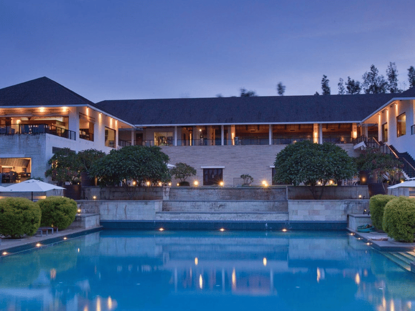 Evening view of The Serai Chikmagalur with illuminated modern villa, steps leading to a pool, poolside loungers, umbrellas, and neatly trimmed greenery.