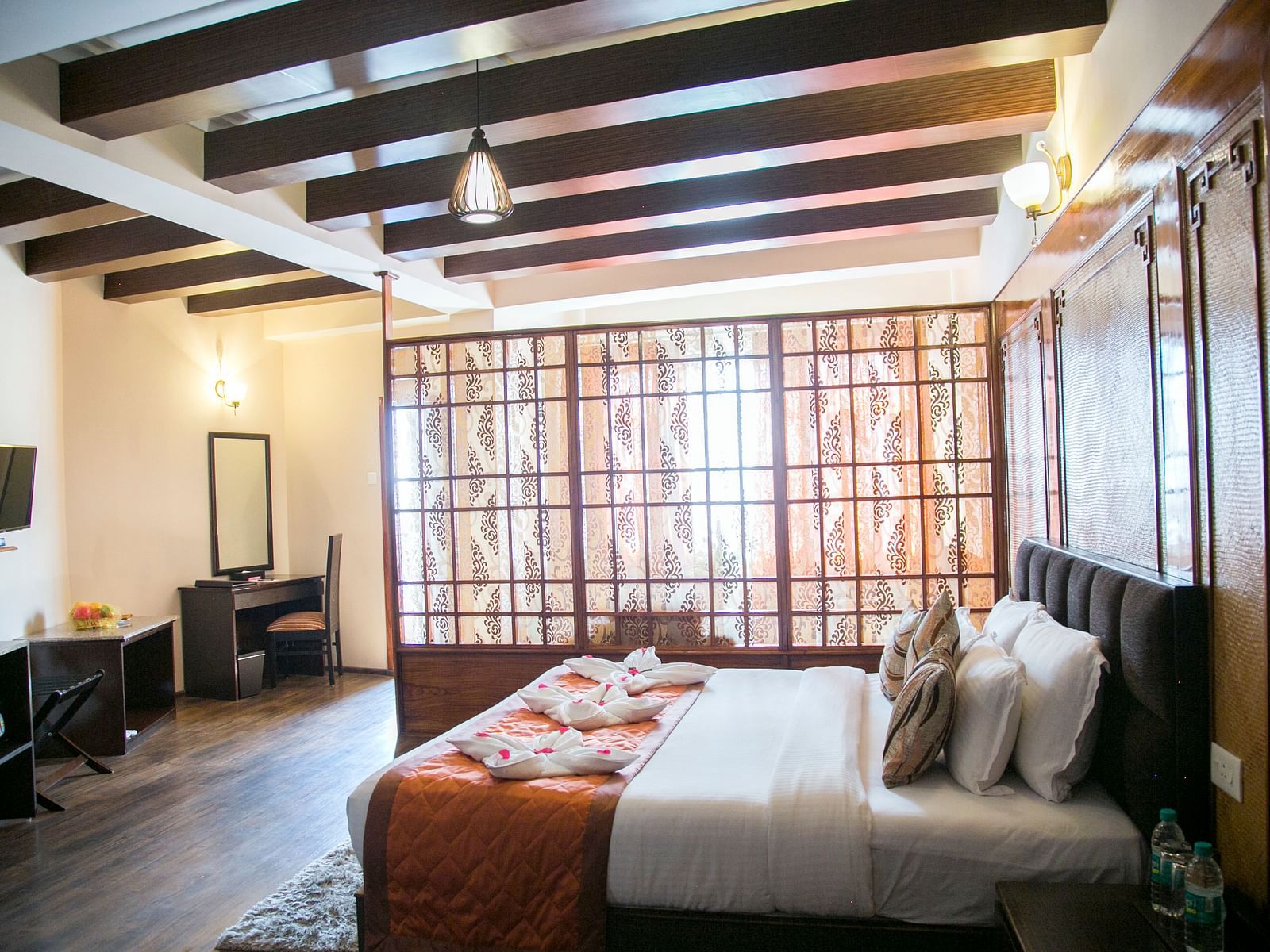 A room at New Orchid Lords Inn Gangtok contains a bed with pillows and towel swans, a bedside table with water bottles, a desk with a mirror and chair, and a TV.