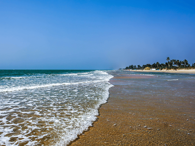 A view of Varca Beach, one of the most popular beaches in South Goa, with the foamy waves crashing on the shoreline with trees in the distance and blue sky above.