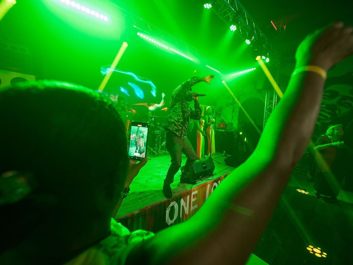 A person with their arm raised is silhouetted against bright  laser lights in a dimly lit club. The person's back is to the viewer, and the scene is energetic and dynamic.