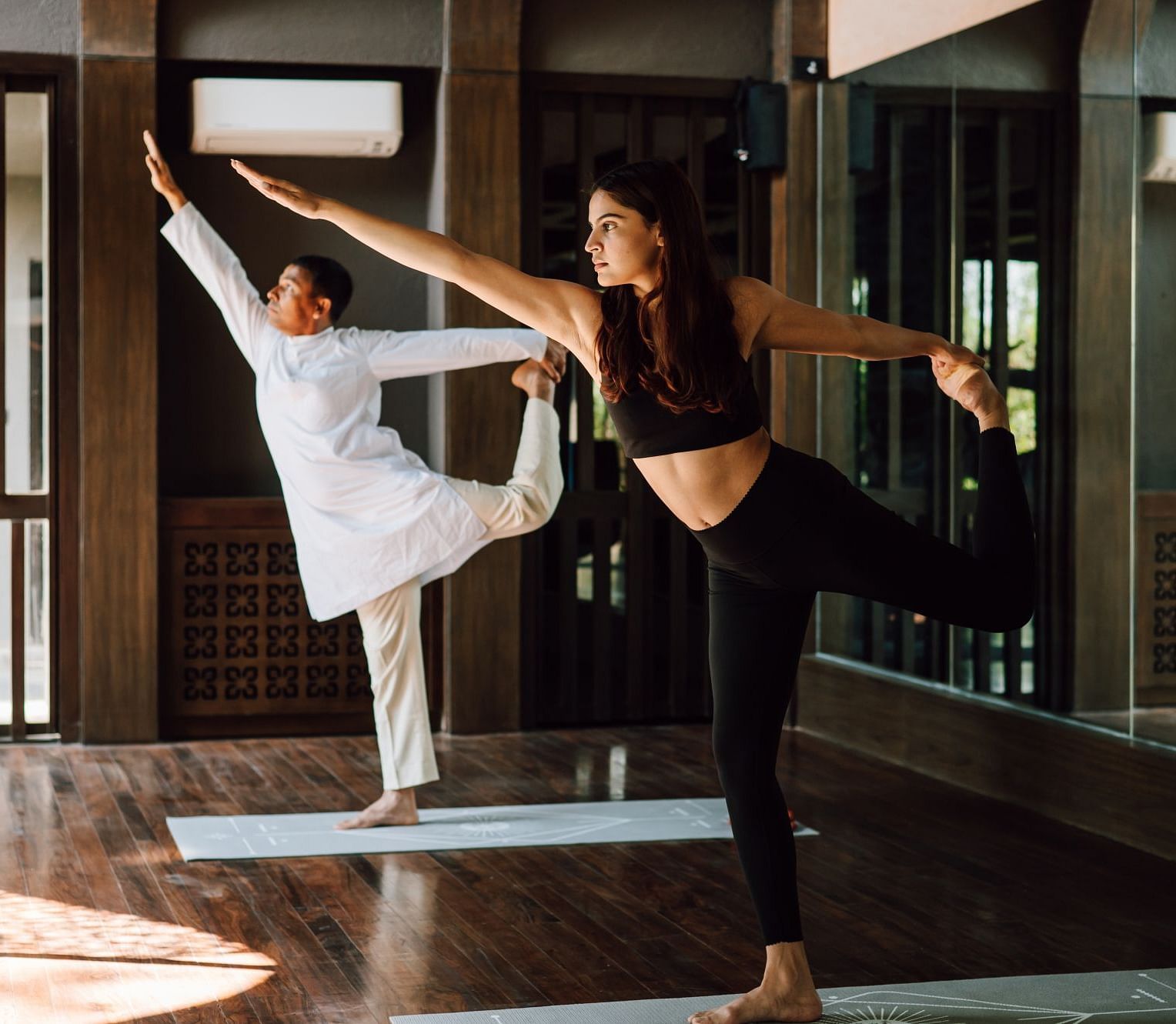 Two individuals practicing yoga indoors, performing balance poses with focused expressions.