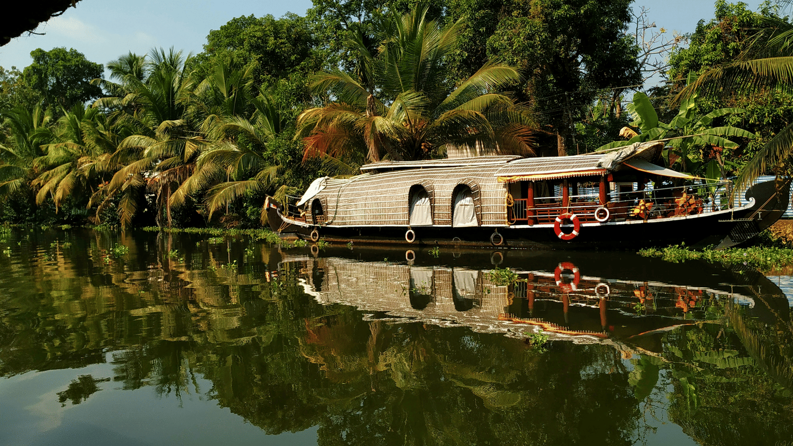 A traditional houseboat with a thatched roof is floating on the calm backwaters, flanked by lush trees reflecting on the water