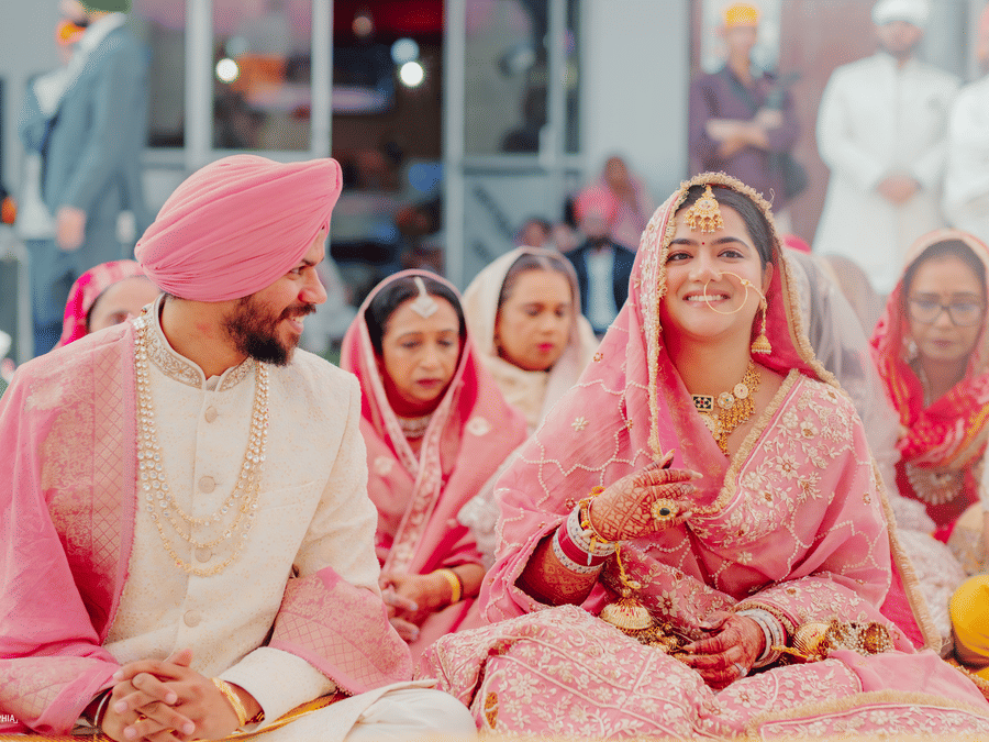 A smiling bride in a detailed pink lehenga sits next to her groom in a pink turban and cream sherwani during a traditional wedding ceremony at Umaid Palace.