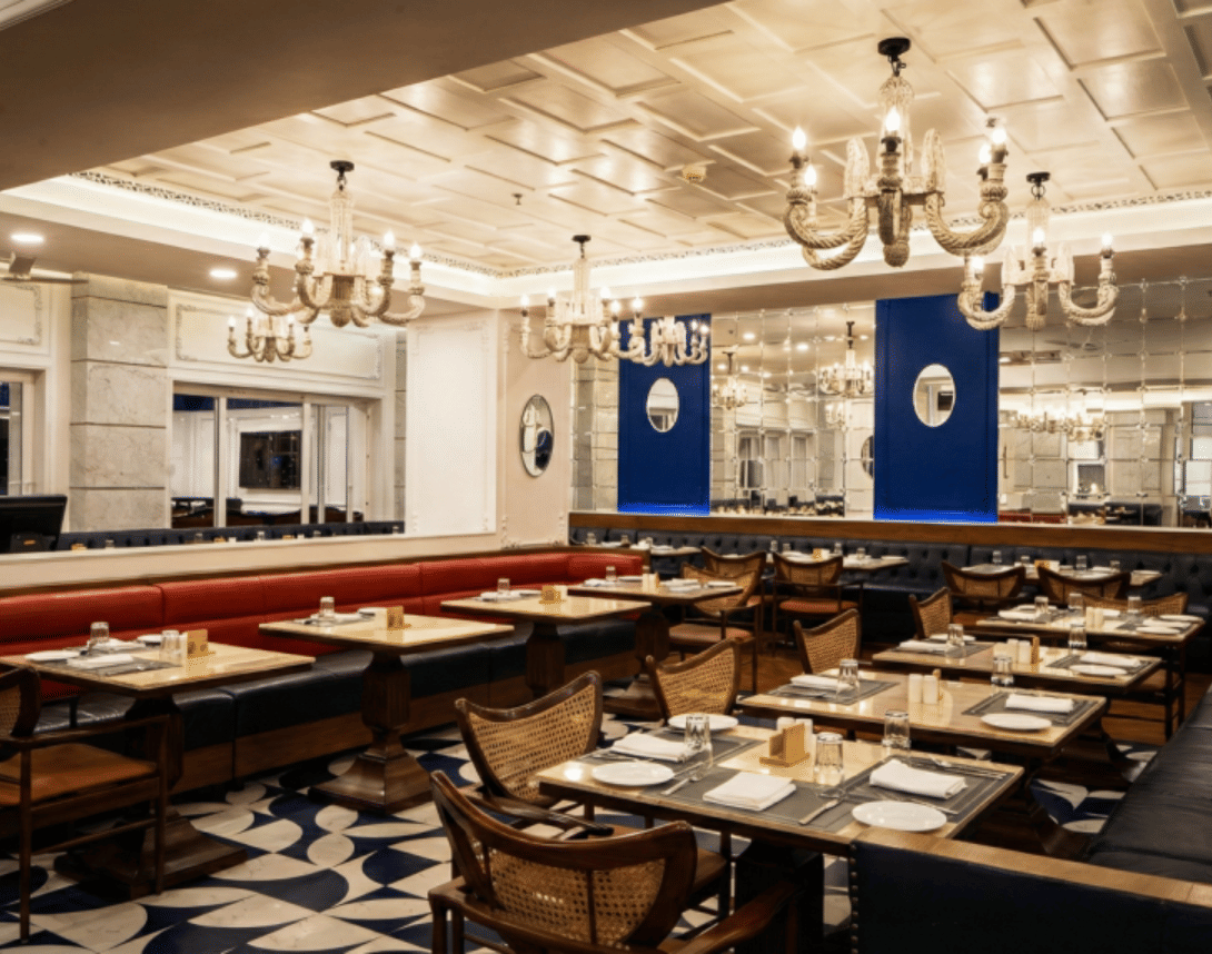 An outdoor dining or lounge area with square patterned pavement, trees wrapped in lights, small tables, chairs, and large red umbrellas under a sunset or twilight sky.